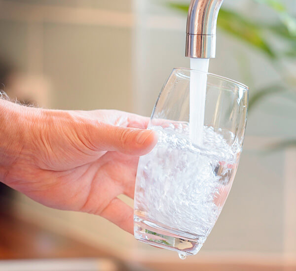 Käsi pitelee vesilasia juoksevan veden alla kuvaten toimivan vedenjakelun tärkeyttä.