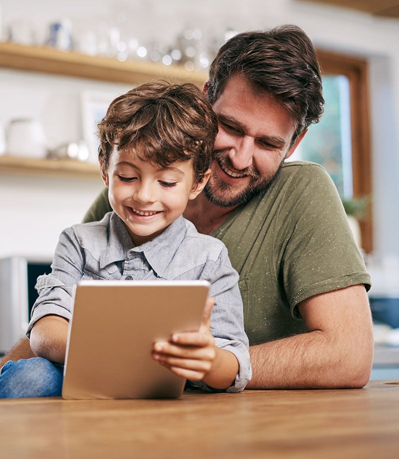 Father and son are smilingly looking at a tablet by a table.