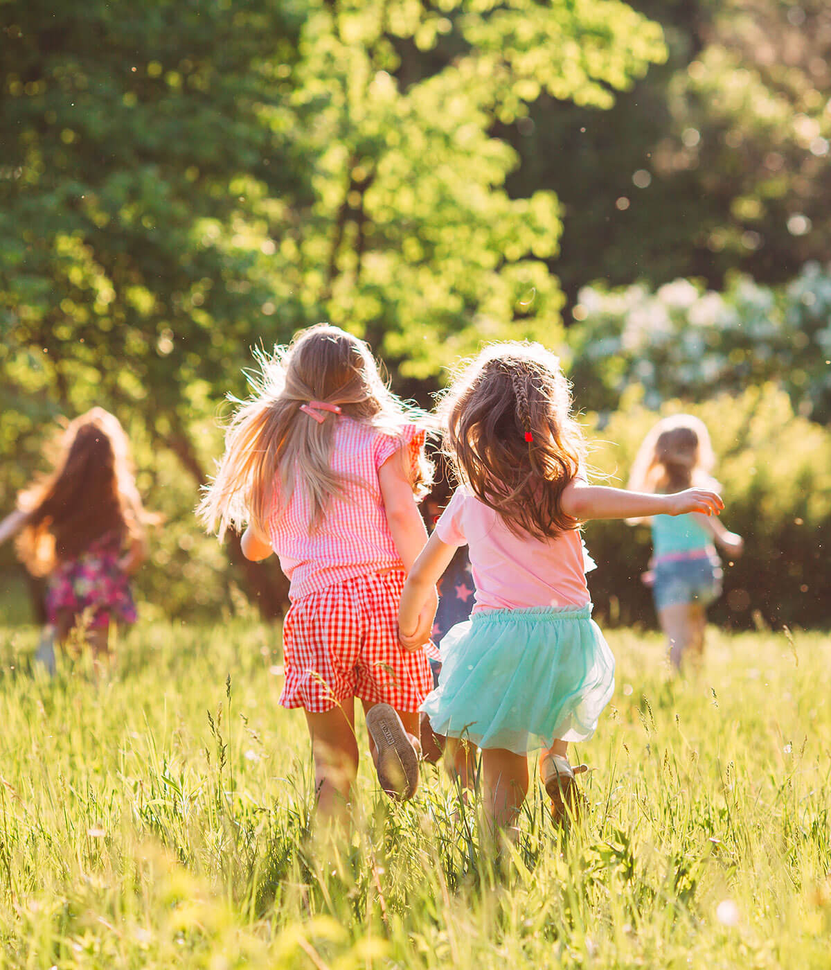 A group of young girls running through a sunny green meadow toward a tree line, seen from behind, wearing colorful summer clothing.