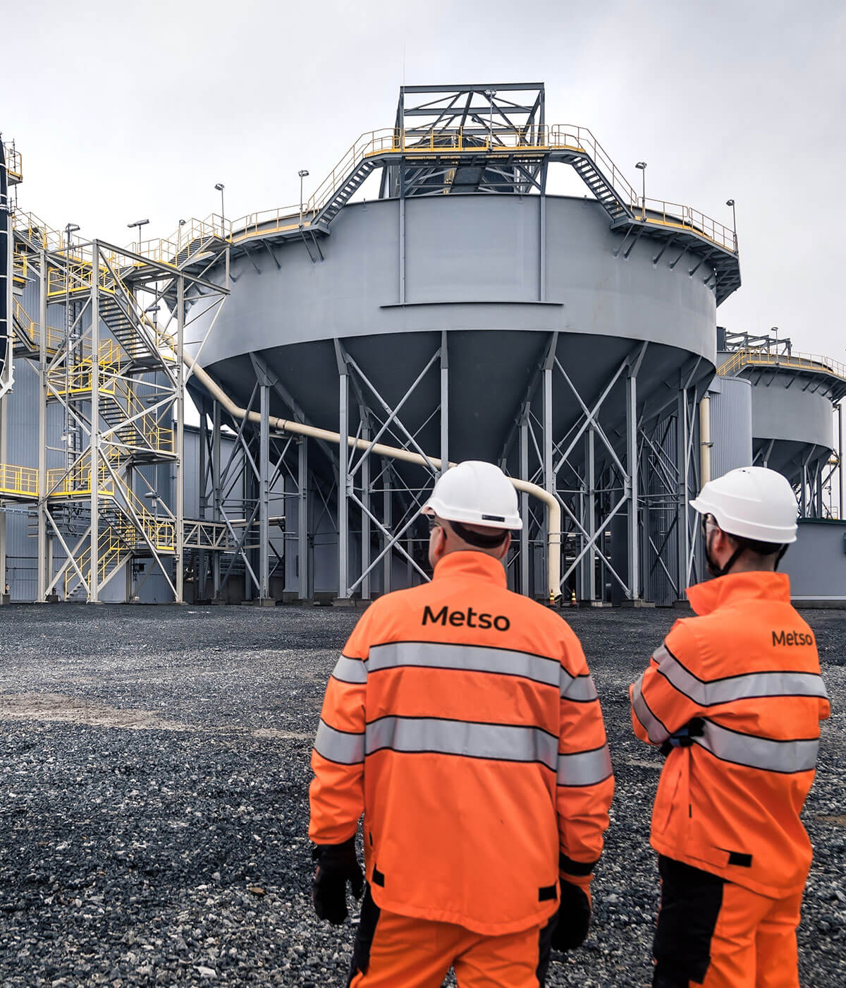 Two Metso workers in orange high-visibility jackets and white hard hats standing at an industrial facility, looking up at large grey storage tanks.