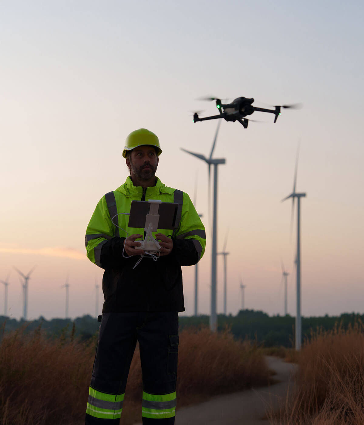 A worker in a yellow high-visibility jacket and hard hat operating a drone at a wind farm at dusk, with multiple wind turbines visible in the background.