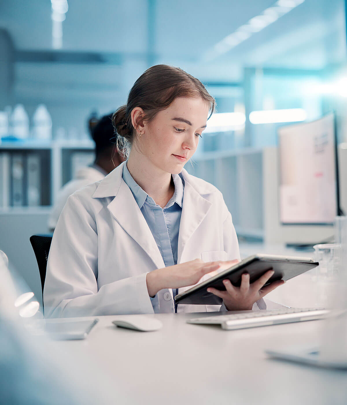 A young woman in a white lab coat using a tablet at a desk in a modern laboratory environment.