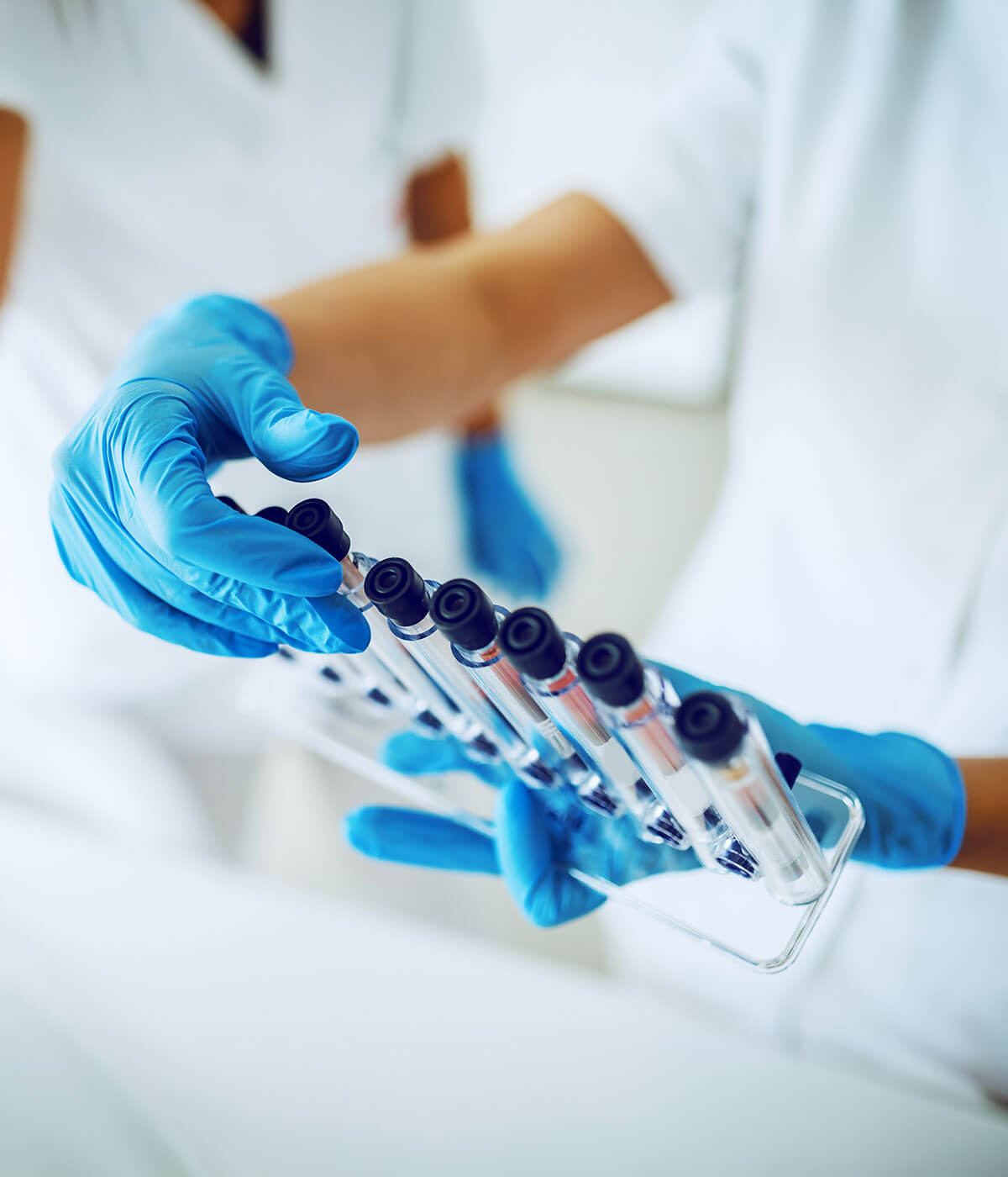 A lab worker in blue gloves and a white coat handling a rack of sealed sample vials in a clinical setting.
