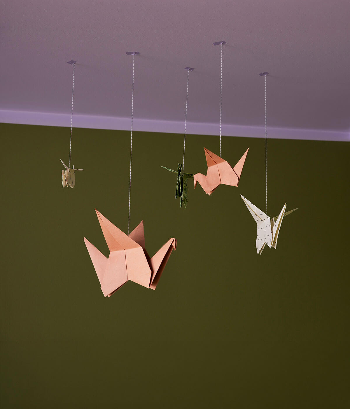 Five origami paper cranes in peach, green, white, and cream tones hanging from strings against an olive green wall with a lilac ceiling.