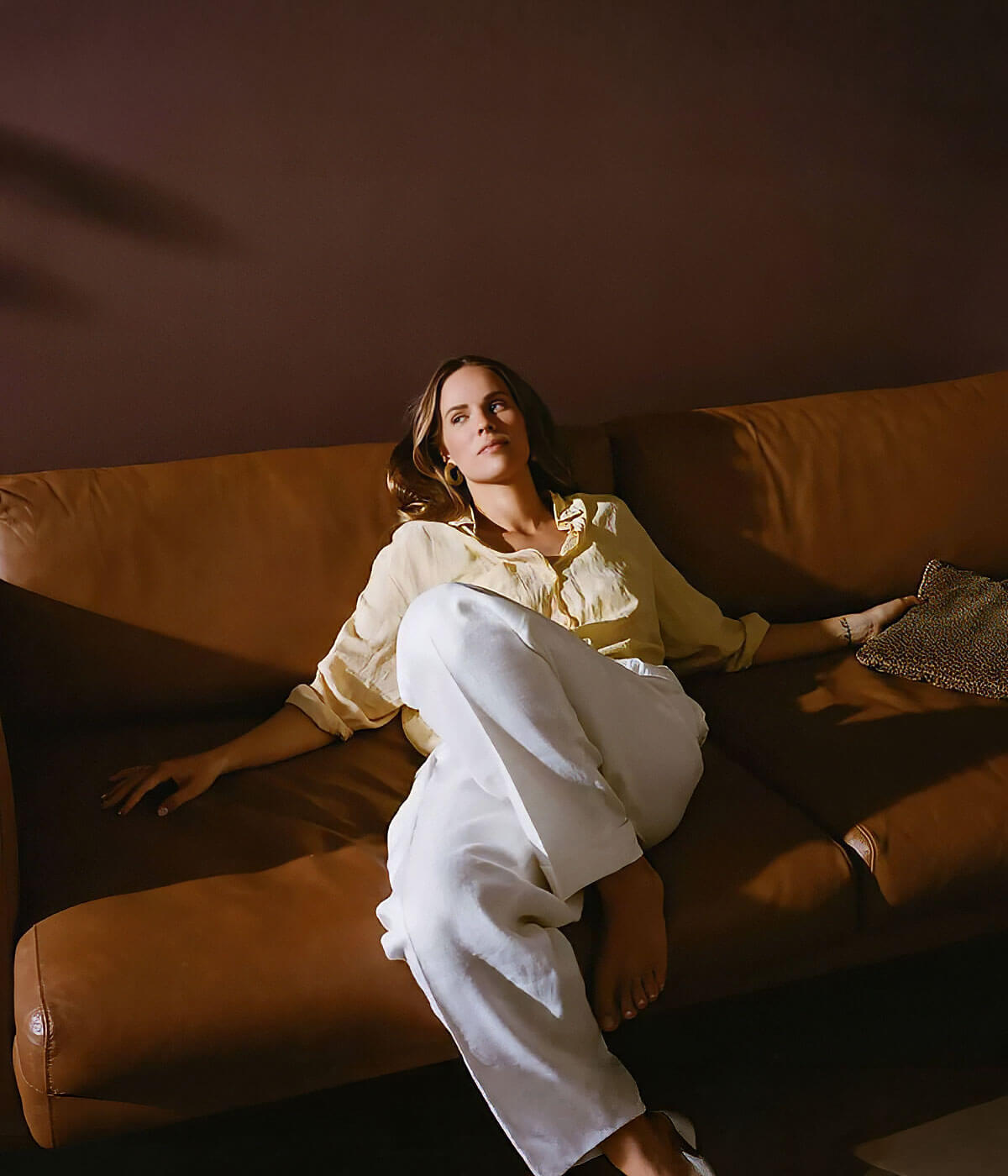 A woman in a cream blouse and white trousers reclining on a tan leather sofa against a warm dark brown wall, lit by soft natural light.