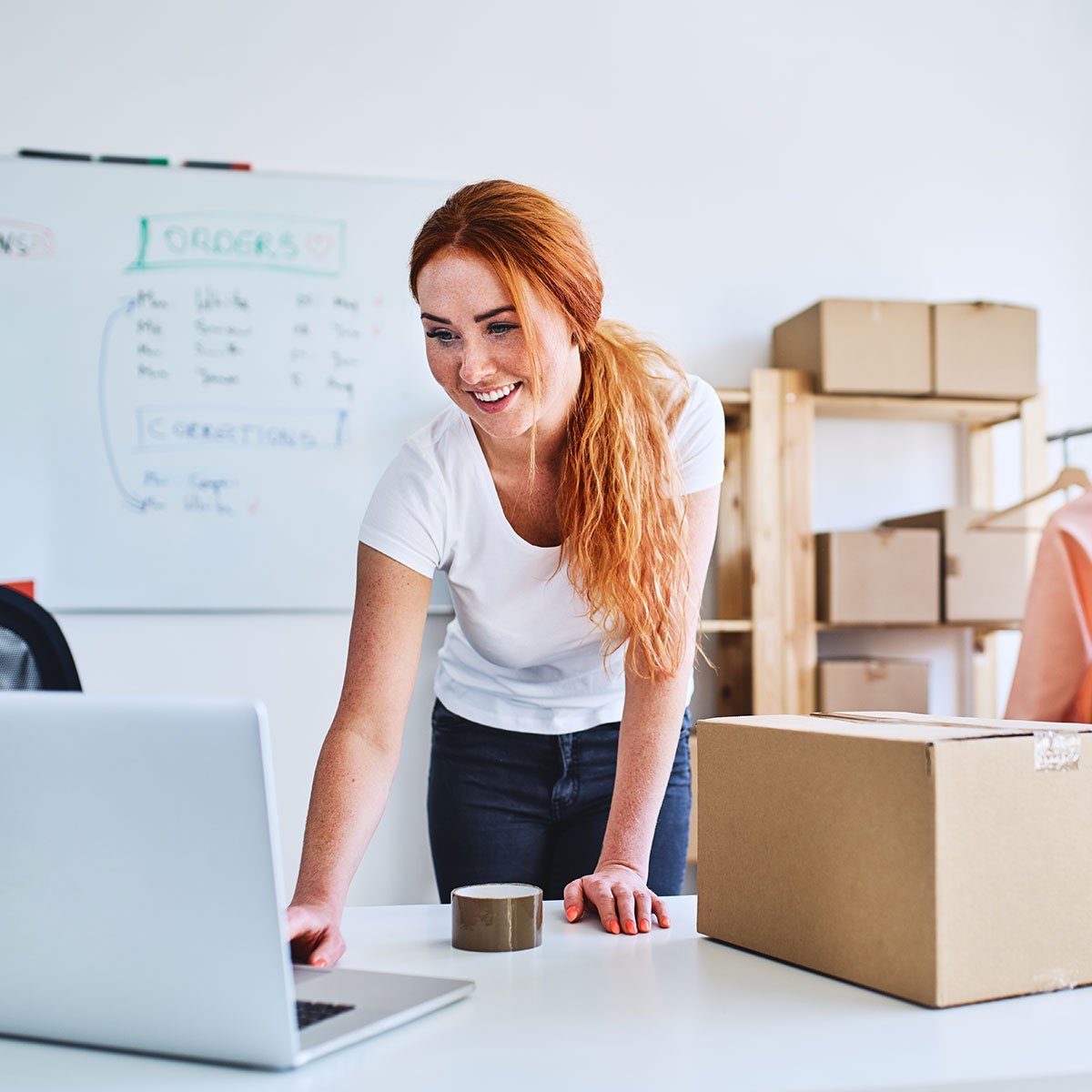 A cheerful red-haired woman in a white t-shirt is packing an order from the Adobe Commerce e-commerce store