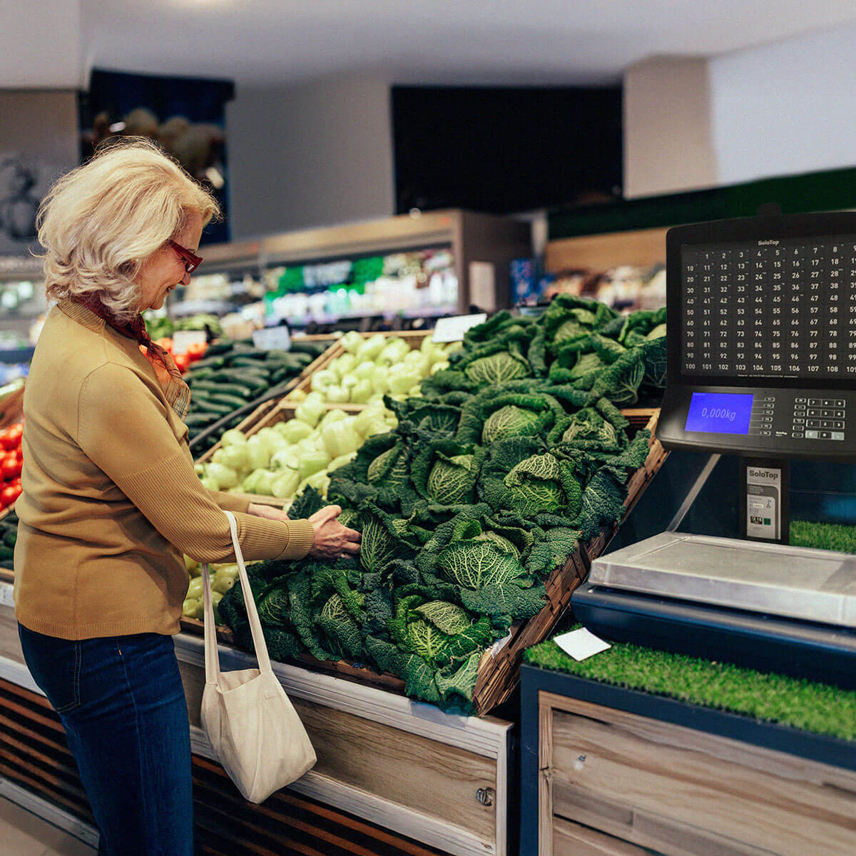 Nainen beigessä neuleessa tarkastelee kaalia ruokakaupan hedelmä- ja vihannesosastolla, vieressä kassajärjestelmään integroitu vaaka