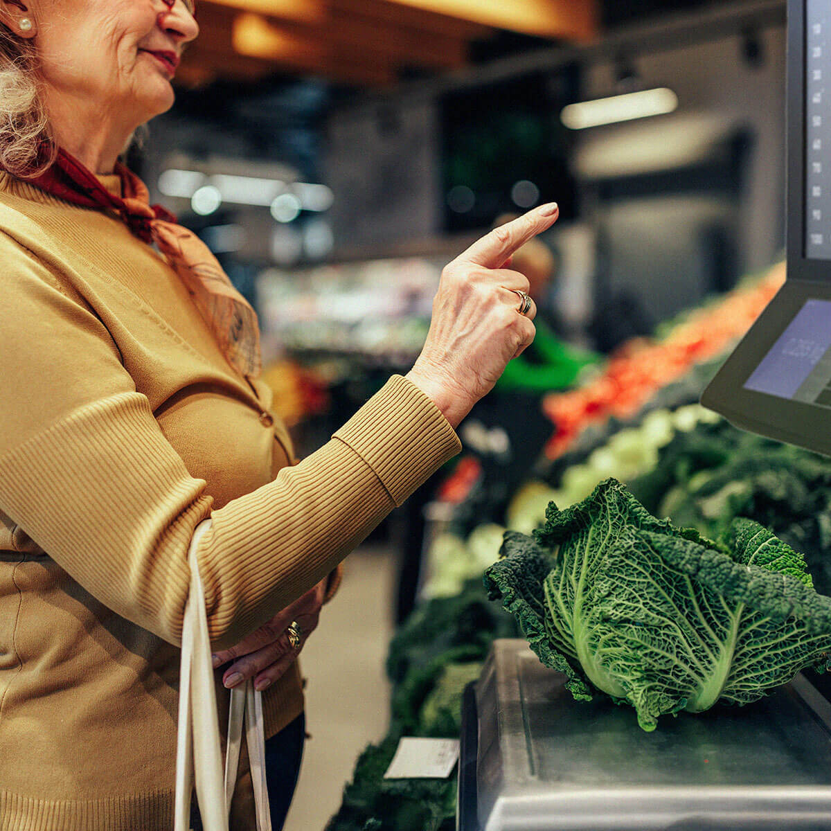 Nainen beigessä neuleessa punnitsee kaalia ruokakaupan hedelmä- ja vihannesosastolla, vieressä kassajärjestelmään integroitu vaaka
