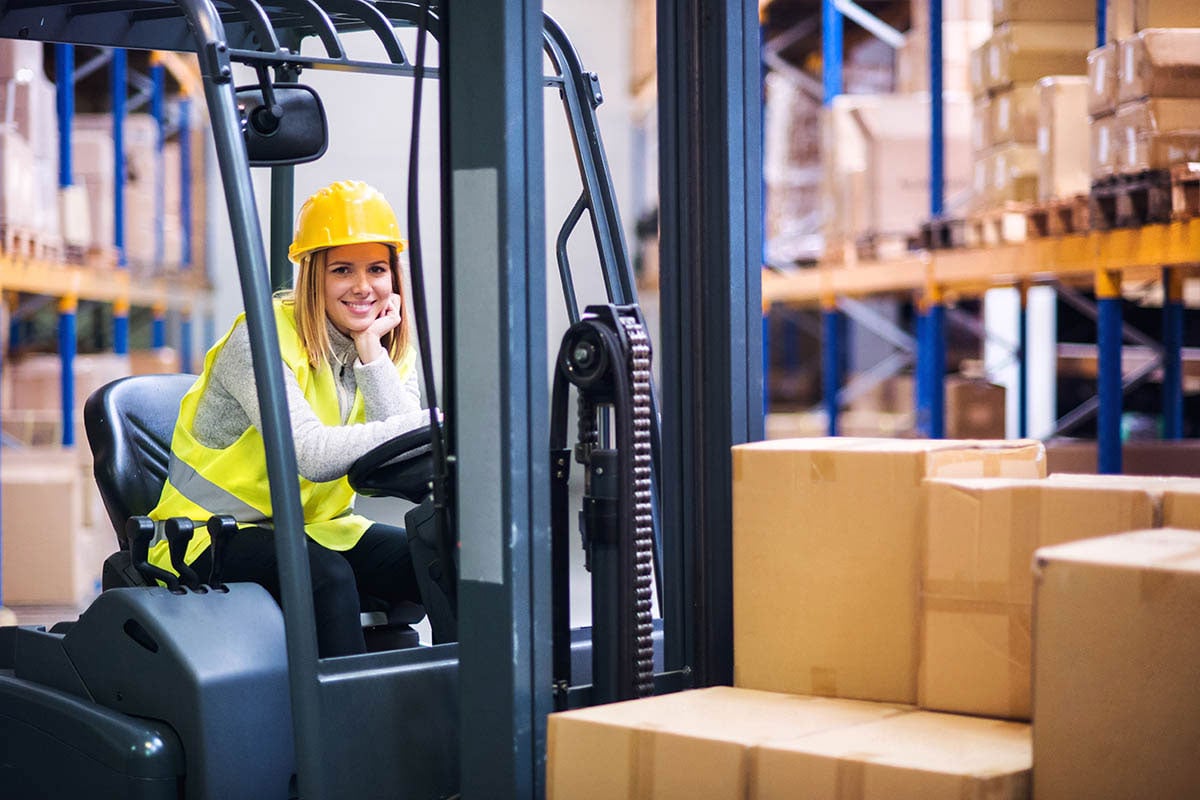 A smiling woman in a yellow vest and helmet leans behind the wheel of a truck in a warehouse
