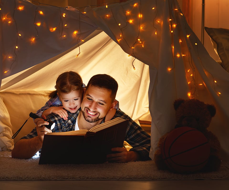 Isä lukee tyttärensä kanssa kirjaa taskulampun valossa teltassa.