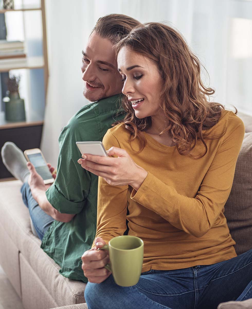  A smiling couple is looking at their mobile phones  on a sofa.