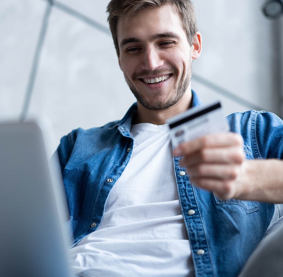 Hymyilevä mies sinisessä farkkupaidassa katsoo maksukortti tehdessään verkkokauppaostoa
