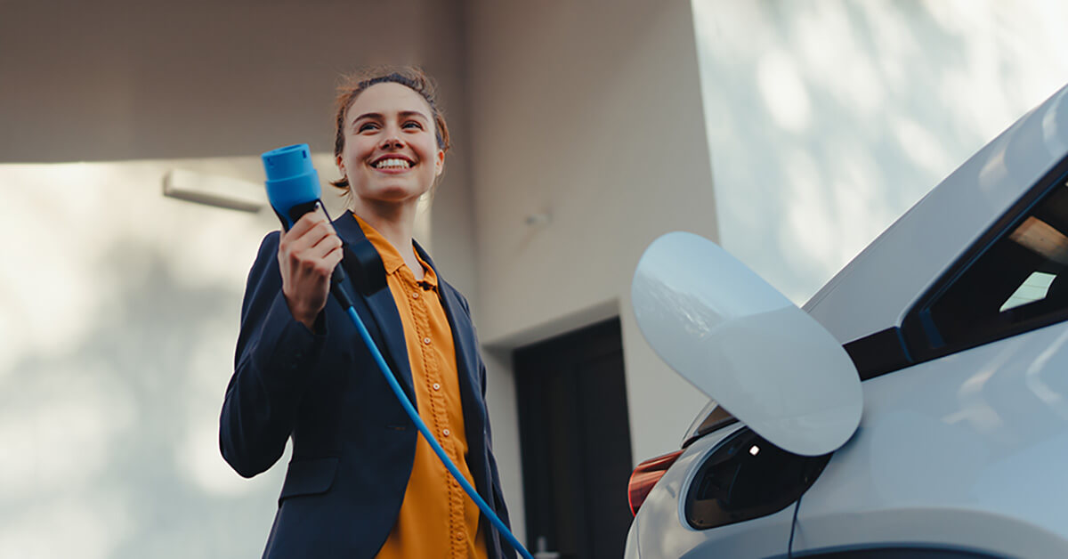 Nainen laittaa sähköauton lataukseen omakotitalon pihalla.