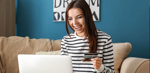 Tummahiuksinen iloinen nainen tekee verkkokauppaostoa dynaamisen hinnoittelun pohjalta kannettavan tietokoneen edessä ja maksukortti kädessä