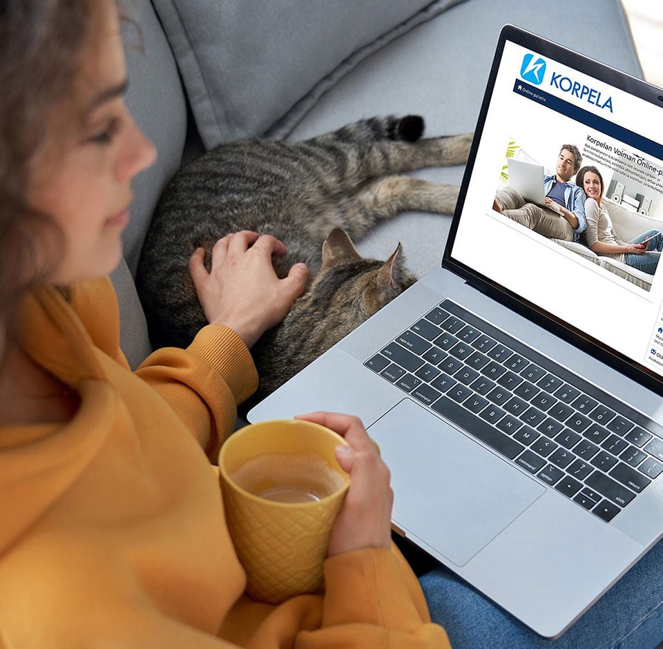 Nainen istuu sohvalla kahvimukin ja läppärin kanssa ja katsoo Korpelan Voiman online-palvelusivuja
