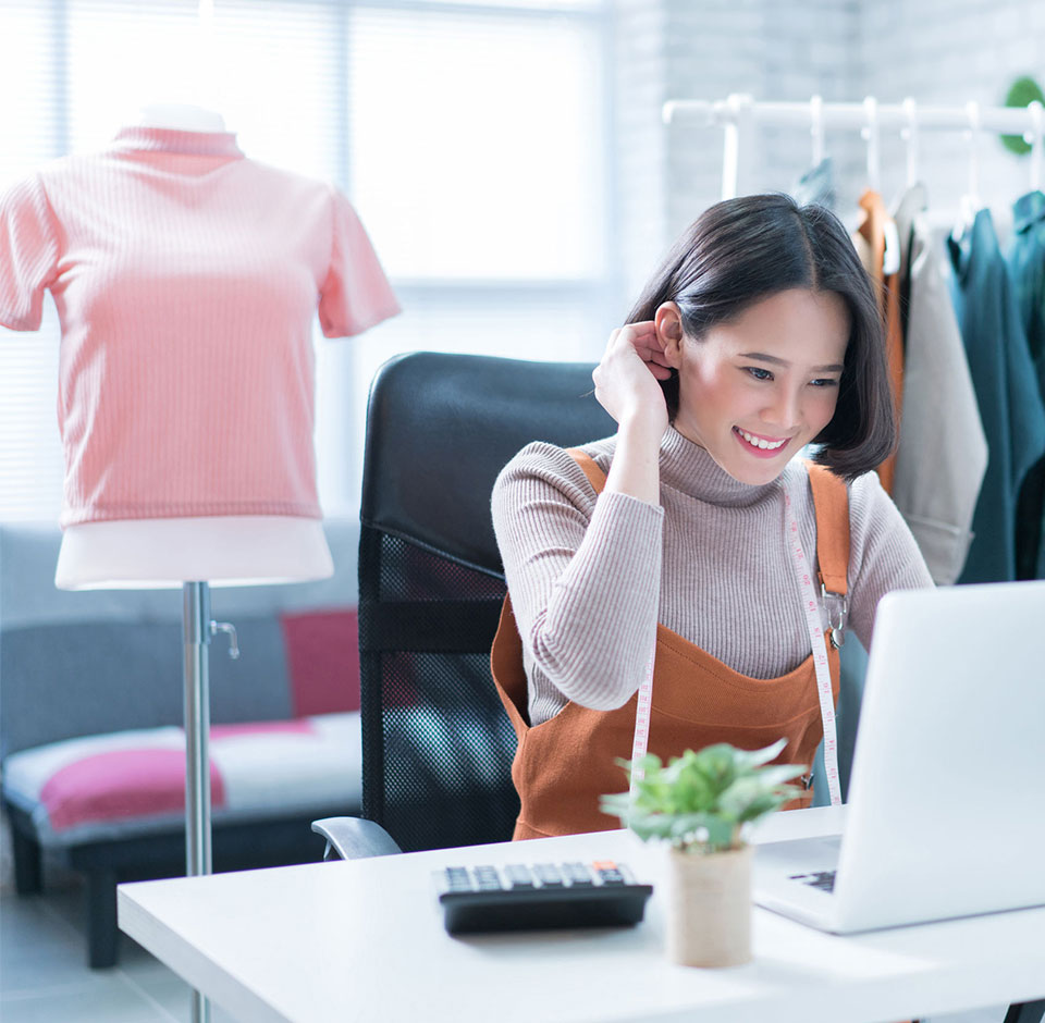 Hymyilevä vaatemyymälän verkkokauppias katsoo kannettavan tietokoneen näytöltä WooCommerce verkkokauppansa kassaintegraation tietoja, taustalla vaatekaupan vaatteita rekissä ja vaaleanpunainen neulepaita sovitusnuken päällä