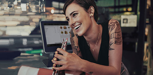 Solteq Commerce Cloud - Mørkhåret smilende servitør ser på kameraet og i bakgrunnen kan du se en elegant restaurant med Solteq Cloud POS-system