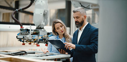Two professionals reviewing data on a clipboard next to an industrial robot arm on a manufacturing floor