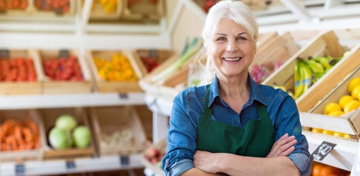 Hymyilevä nainen kauppansa edustalla jonka toimitusketjun ohjaus on kunnossa, taustalla kasviksia ja hedelmiä laatikoissa