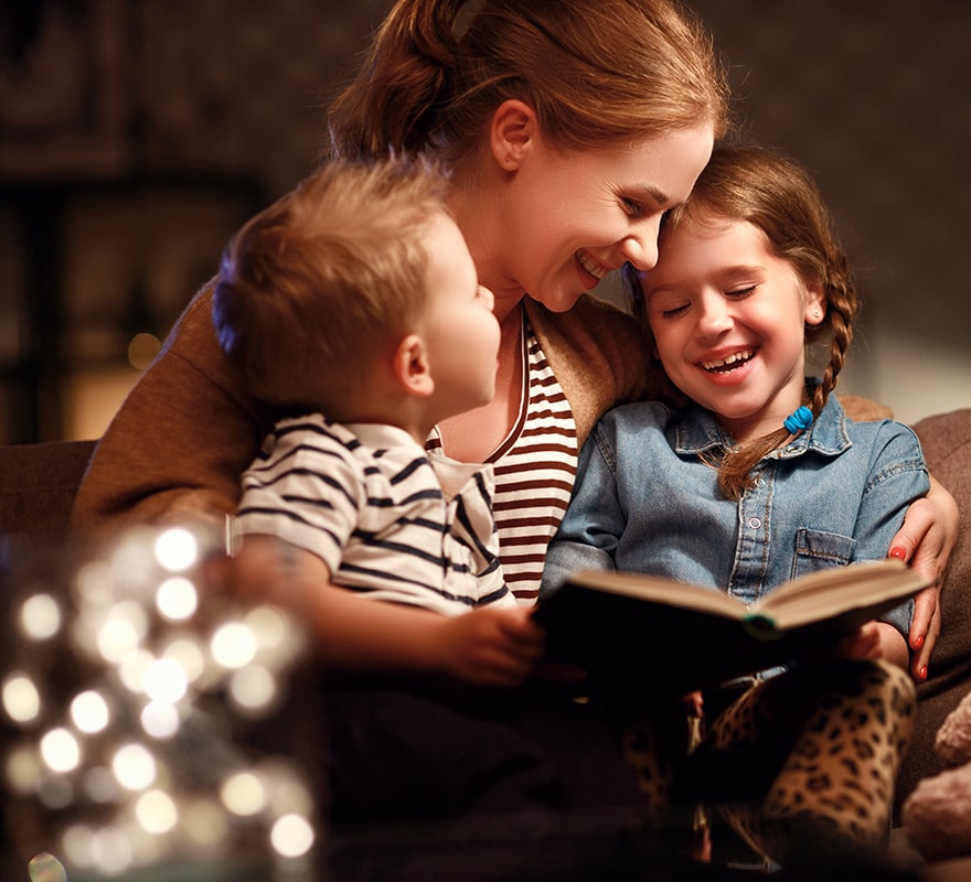 En mamma läser en bok för sina barn på en mysig soffa hemma.