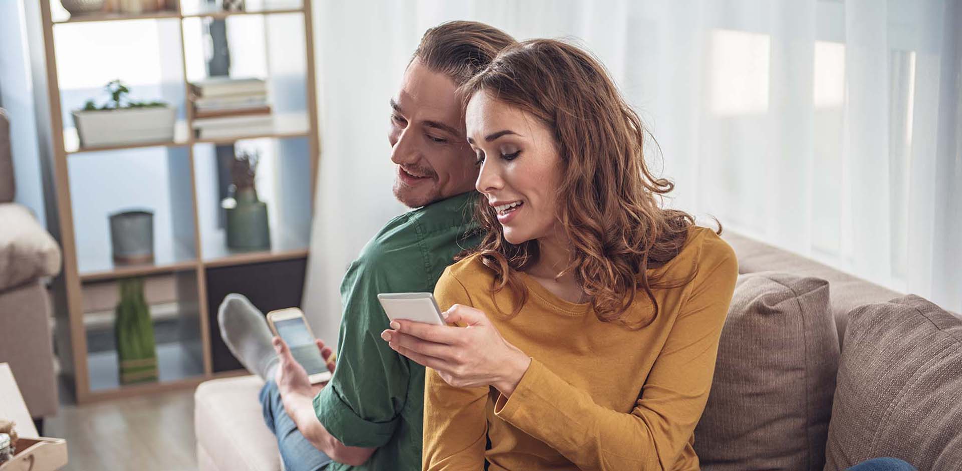  A smiling couple is looking at their mobile phones  on a sofa.